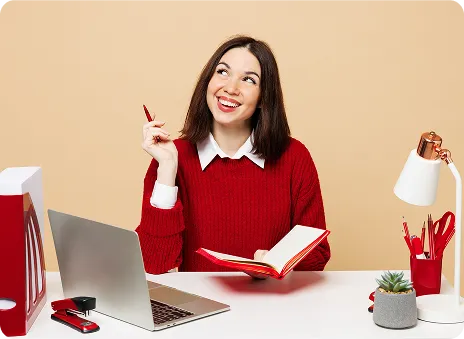 Young woman in a red sweater holding a notebook, sitting at a desk with a laptop and office supplies.