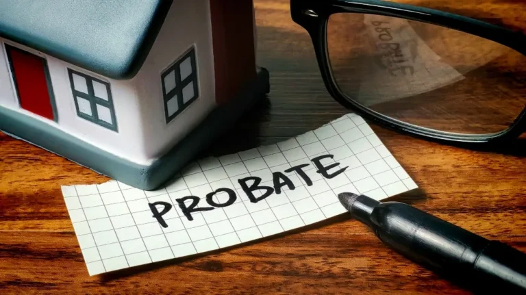 Small gray and white house model next to glasses and a marker on wooden surface with "PROBATE" written on a grid paper.