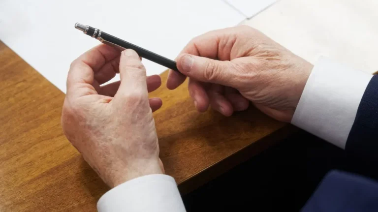 Close-up of elderly hands holding a black pen over a wooden desk with white paper.
