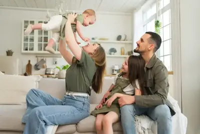 A family of four in a bright living room, mother lifting baby, father and daughter seated on sofa.