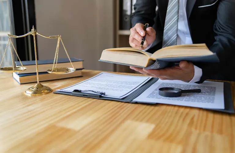 Person in formal attire reviewing a book and legal documents on a wooden desk with scales of justice.