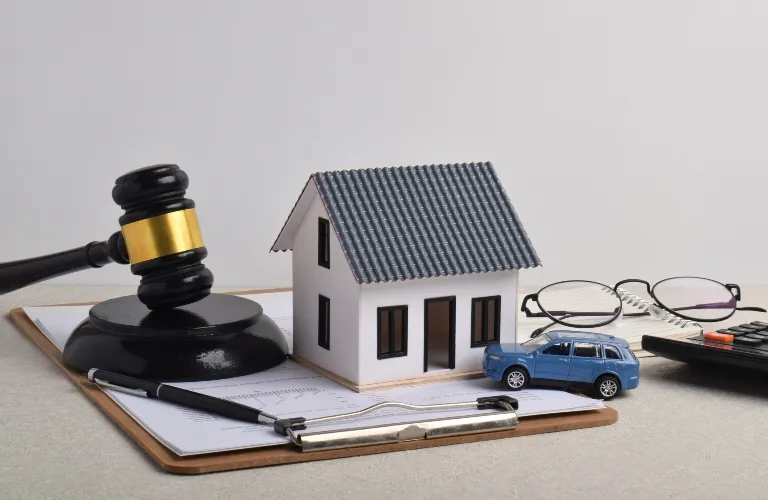 Wooden judge gavel, miniature house model, toy car, clipboard with documents, eyeglasses, and calculator on desk.