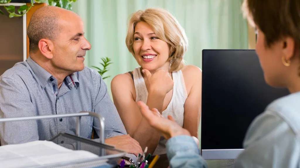 Two middle-aged individuals engaged in conversation with a professional at a desk with a computer.