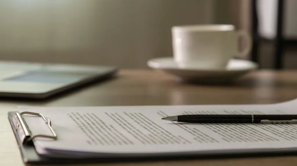 Close-up of a clipboard with documents and a pen on a wooden desk, blurred coffee cup and laptop in background.