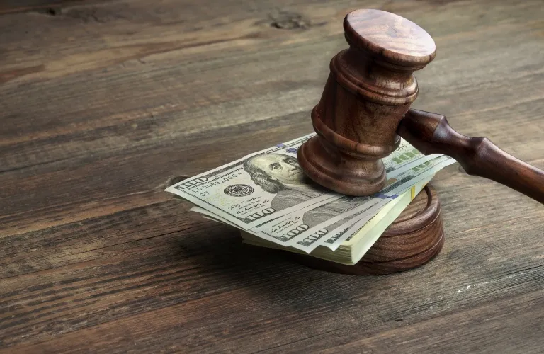 Wooden judge's gavel resting on a stack of U.S. hundred-dollar bills on a wooden surface.