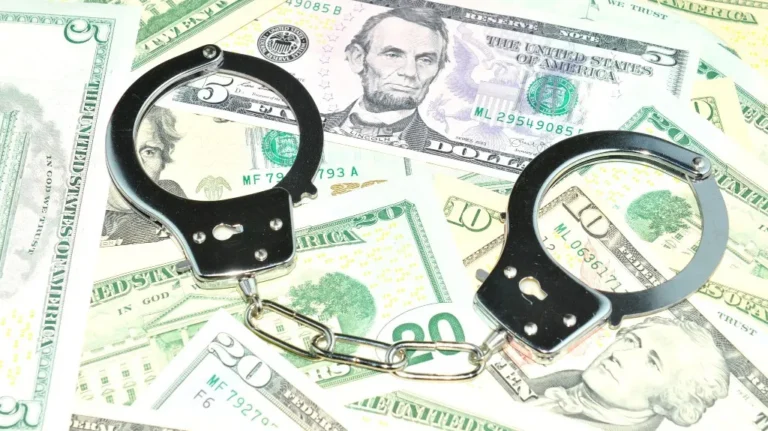 Black metal handcuffs placed on scattered U.S. dollar bills of various denominations.