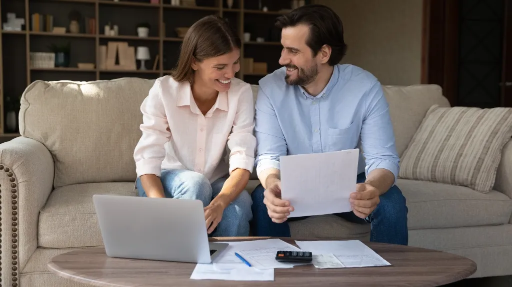 A smiling man and woman seated on a beige sofa reviewing documents with a laptop on a wooden table.