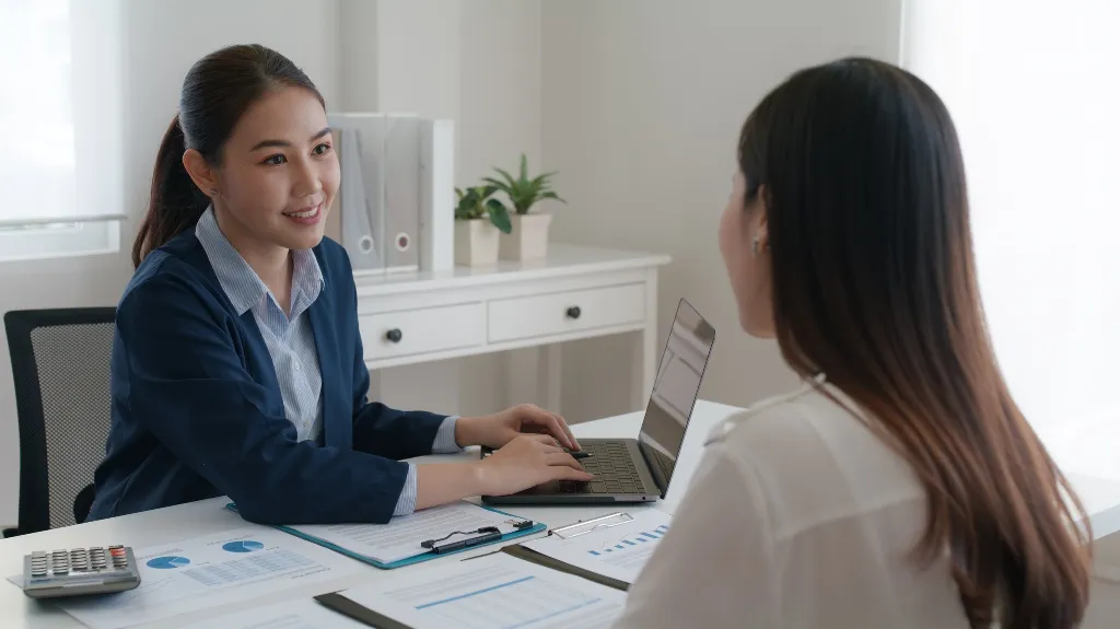 Two professional women engaged in a discussion with documents and a laptop on a white office desk.